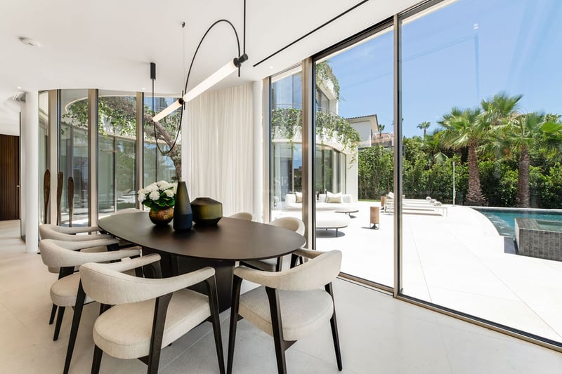Salle à manger intérieure avec table en bois sombre et chaises beiges donnant sur une terrasse ensoleillée avec piscine.