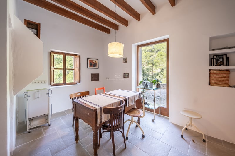 Salle à manger lumineuse avec table en bois, poutres apparentes et porte vitrée donnant sur la forêt.