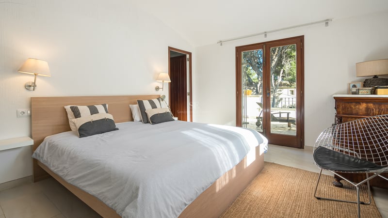 Chambre lumineuse avec lit en bois, murs blancs et portes vitrées ouvrant sur une terrasse ensoleillée.