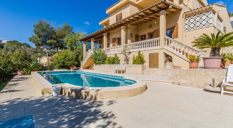 Grande piscine privée devant une villa méditerranéenne ocre avec terrasse et collines verdoyantes.