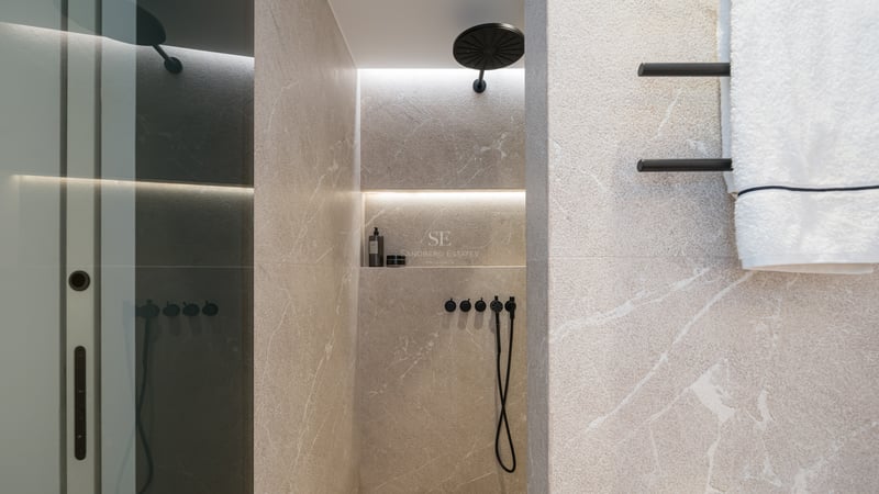 Douche avec carrelage en pierre beige, pommeau de pluie noir mat et niche éclairée sur un mur en pierre veinée.