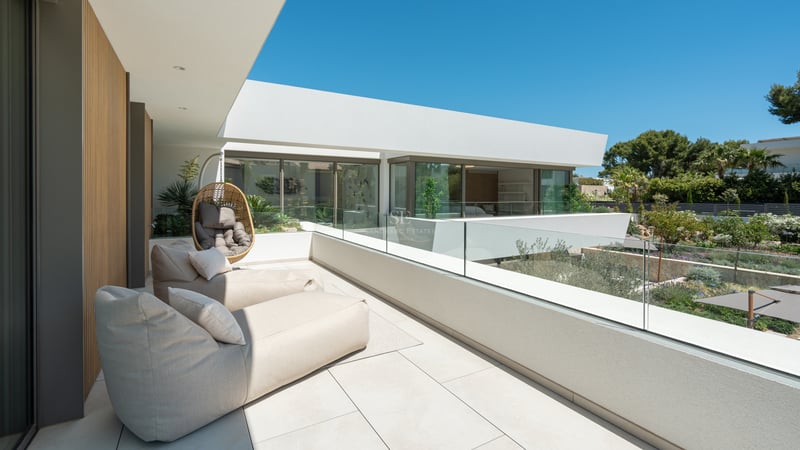 Vaste terrasse blanche avec chaises longues beiges, garde-corps en verre et vue sur un jardin paysager sous un ciel bleu.