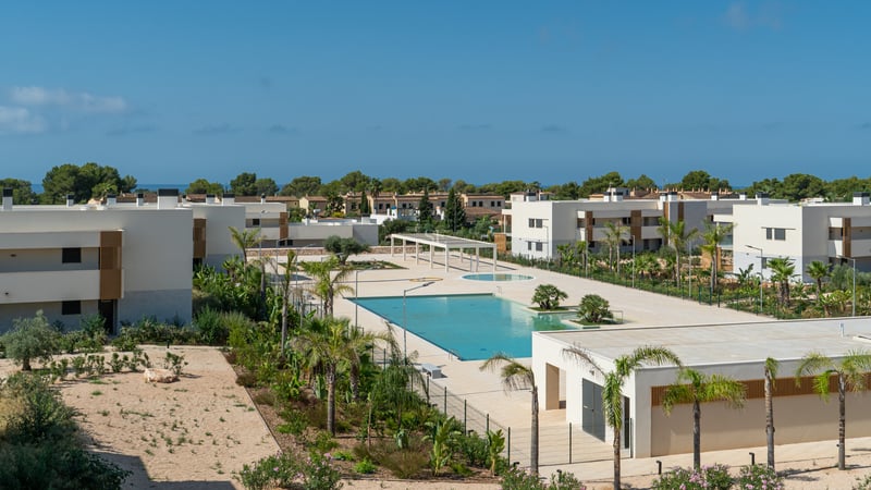 Vista elevada de una gran piscina turquesa rodeada de edificios modernos blancos y palmeras bajo un cielo azul.