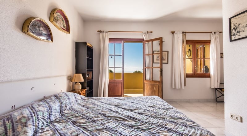 Chambre lumineuse avec des portes-fenêtres en bois ouvrant sur une vue mer, des murs blancs et une décoration traditionnelle.