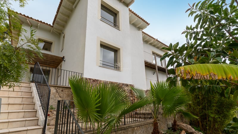 Façade de villa blanche avec soubassement en pierre, escaliers extérieurs et palmiers sous un ciel dégagé.