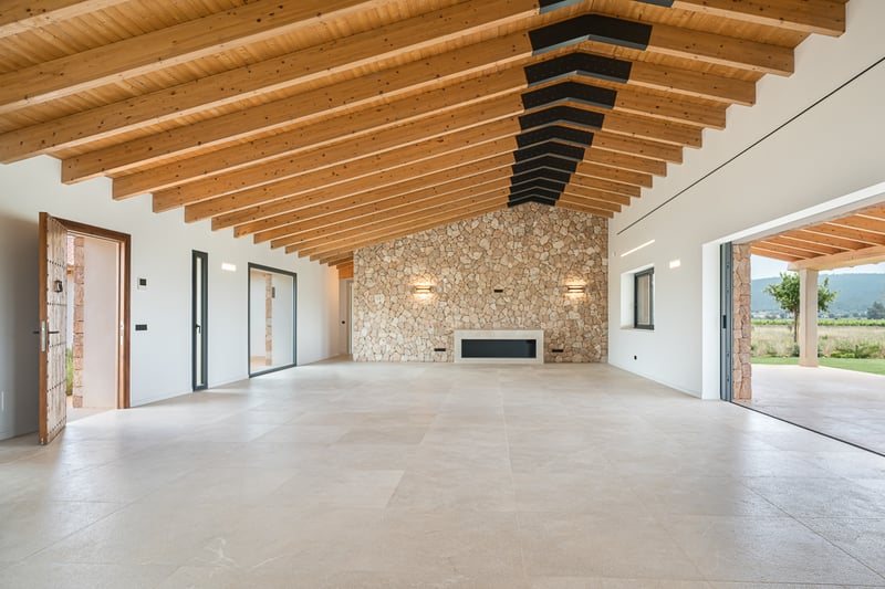 Grand salon avec hauts plafonds à poutres apparentes, mur en pierre naturelle et baies vitrées sur le jardin.