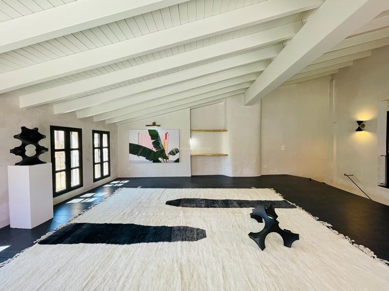 Salon spacieux avec plafond à poutres blanches, sol sombre et grand tapis artisanal aux motifs noirs.