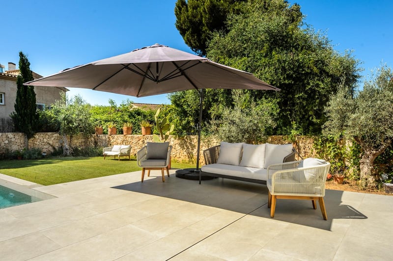 Terrasse ensoleillée avec canapé gris moderne, fauteuils et grand parasol à côté d'une piscine et d'un jardin.
