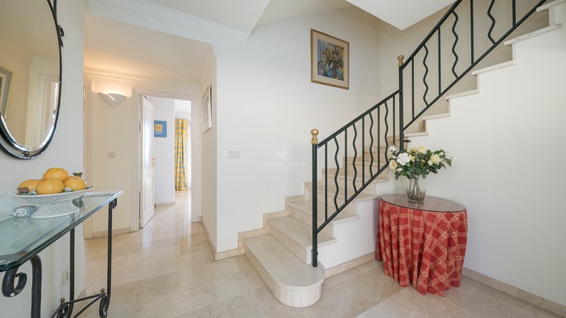 Hall d'entrée lumineux avec escalier en marbre crème, rampe en fer forgé et sol en pierre polie.