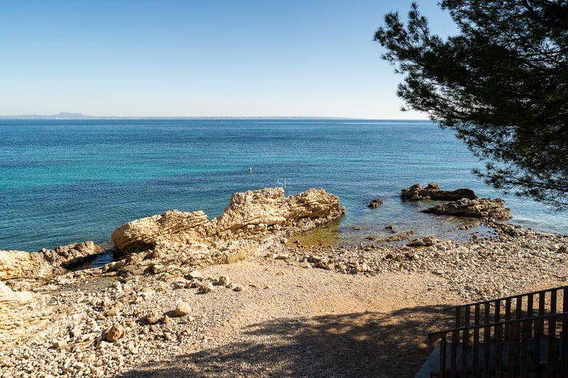 Vue surélevée d'une crique rocheuse méditerranéenne aux eaux turquoises et horizon dégagé sous un ciel bleu.