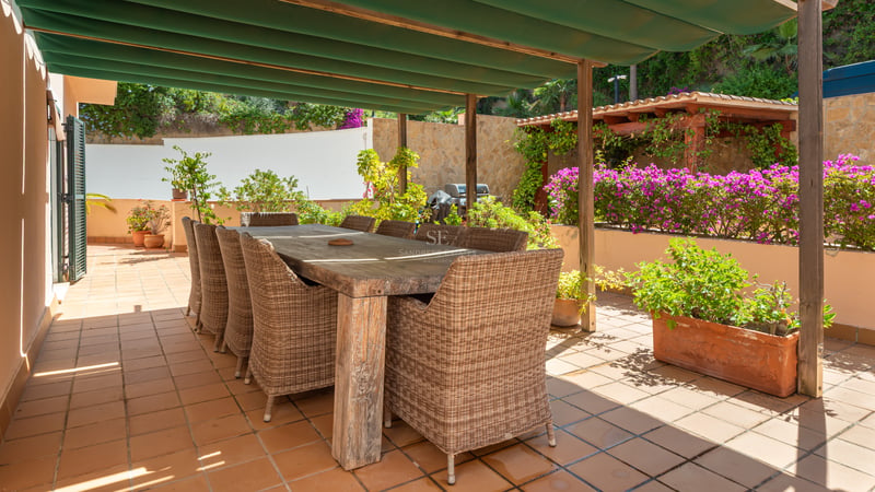 Grande table à manger en bois avec chaises en osier sur une terrasse en terre cuite ensoleillée sous un auvent vert.