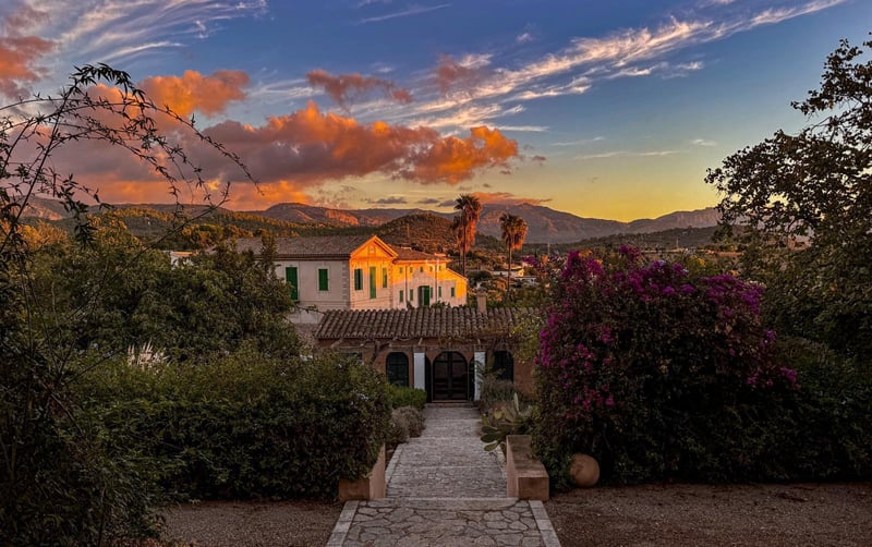Vue panoramique d'une villa méditerranéenne blanche avec toit en terre cuite au coucher du soleil, entourée de jardins et de montagnes.