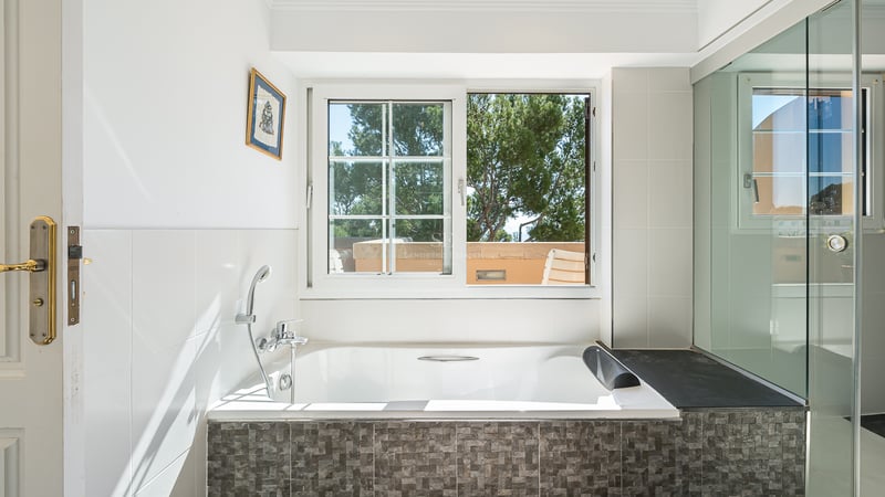 Salle de bain blanche avec baignoire encastrée, mosaïque grise et fenêtre donnant sur des pins méditerranéens.