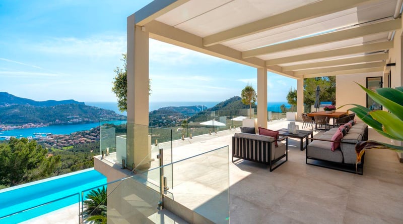 Grande terrasse moderne avec canapés gris, garde-corps en verre et vue imprenable sur une baie et des collines.