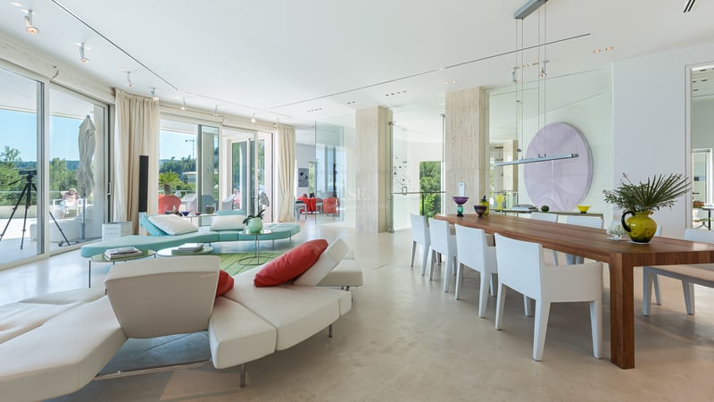 Salon moderne avec sol en pierre, grandes baies vitrées, canapés blancs et table à manger en bois.