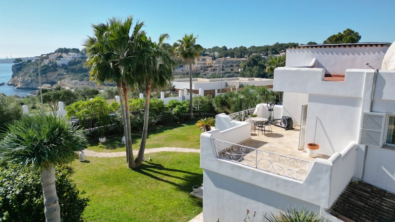 Vue d'en haut d'une villa blanche avec terrasse, jardin avec palmiers et vue sur la mer bleue et les falaises au loin.
