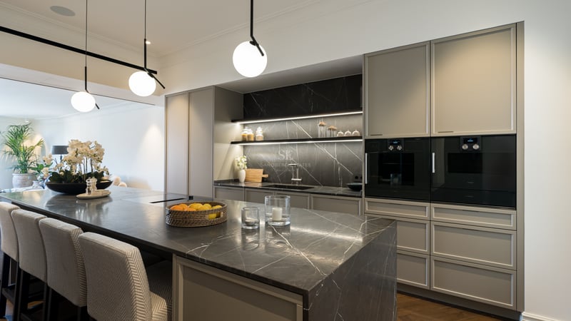 Modern kitchen featuring a dark marble island with seating, grey cabinetry, integrated appliances, and spherical pendant lights.
