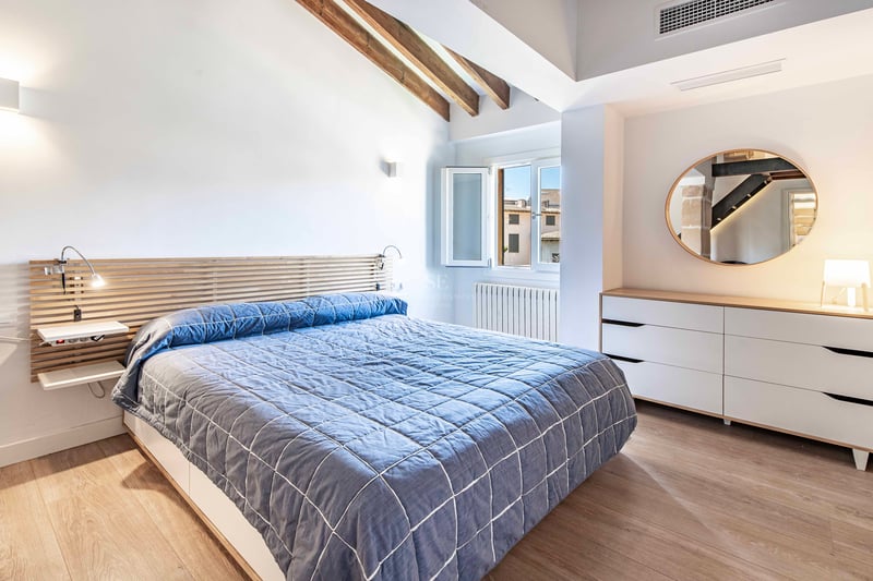 Chambre lumineuse avec murs blancs, poutres apparentes, couvre-lit bleu et un grand miroir rond sur la commode.