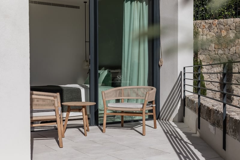 Terrasse moderne avec fauteuils tressés et table en bois, jouxtant une chambre avec grandes baies vitrées.