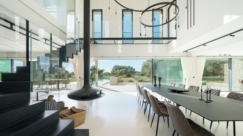 Salle à manger lumineuse à double hauteur avec cheminée suspendue noire, parois vitrées et vue sur le jardin et la piscine.
