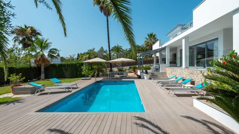 Piscine rectangulaire entourée d'un deck en bois avec transats blancs et palmiers devant une villa moderne.