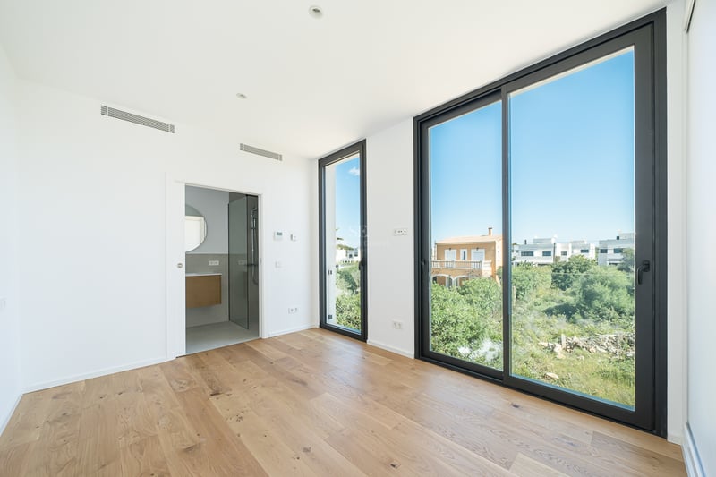 Chambre moderne vide avec parquet clair, grandes baies vitrées noires donnant sur le jardin et salle de bain attenante.
