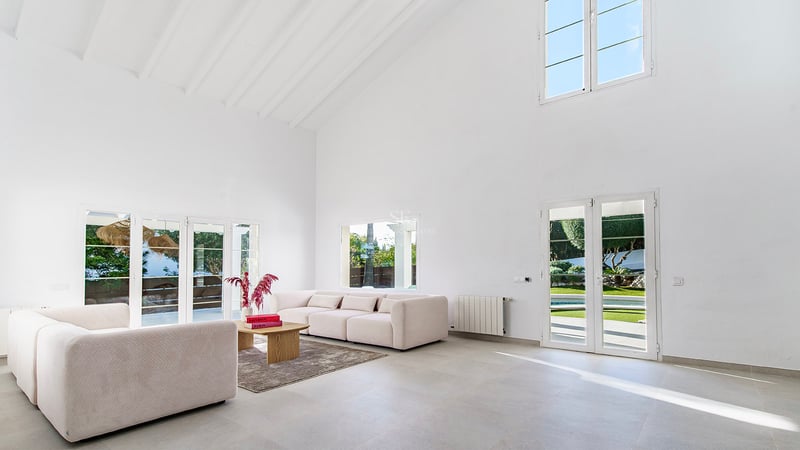 Salon spacieux aux plafonds blancs, canapés beige et portes-fenêtres ouvrant sur le jardin ensoleillé.