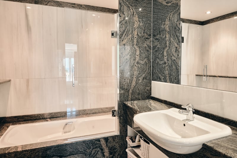 Salle de bain de luxe avec plan de toilette en granit foncé, lavabo blanc et baignoire avec paroi en verre.