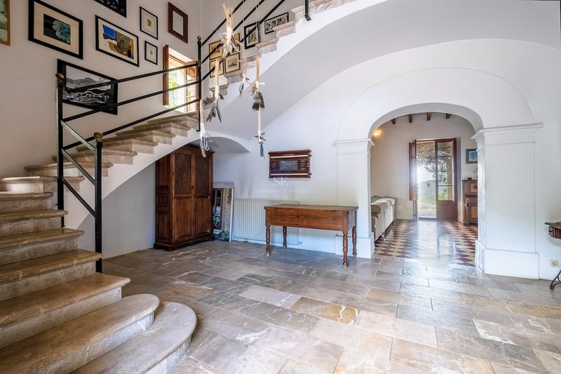 Un grand hall d'entrée avec un escalier en pierre, des meubles anciens en bois et une galerie d'art sous une arche.