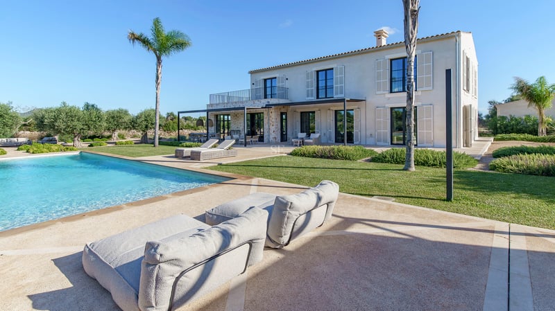 Grande piscine turquoise devant une villa en pierre avec palmiers et pelouse sous un ciel bleu azur.
