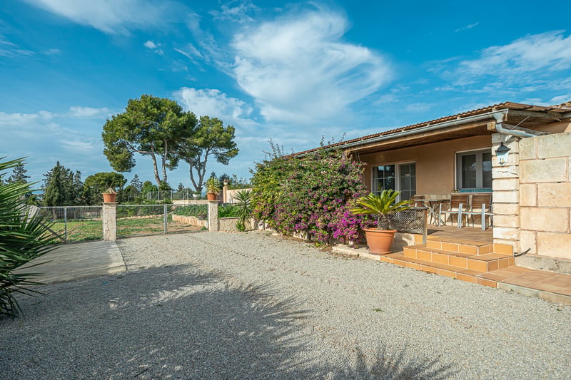 Stenfasad på en villa med terracottaporch, grusplan och bougainvillea under en klarblå himmel.