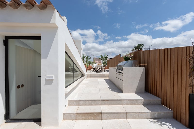 Terraza soleada con cocina de piedra, cerramiento de madera y suelo de piedra caliza bajo un cielo azul.