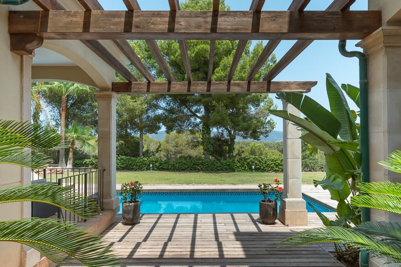 Vue d'une pergola en bois sur une piscine bleue entourée d'un jardin luxuriant et d'arbres méditerranéens.