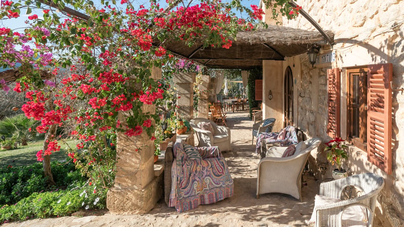 Terrasse en pierre ombragée avec fauteuils en osier, tissus colorés et bougainvilliers roses au soleil.