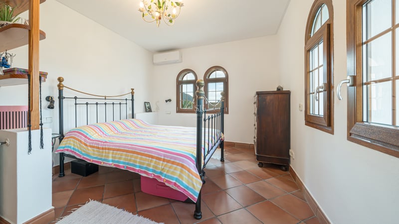Chambre lumineuse avec sol en terre cuite, lit en fer forgé et fenêtres traditionnelles en bois en forme d'arche.