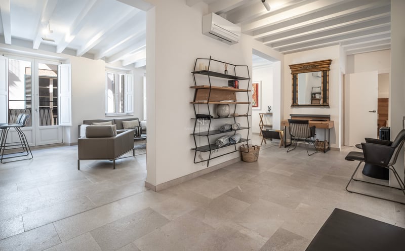 Intérieur moderne avec sol en pierre, poutres blanches, canapé gris et un coin bureau avec miroir doré.