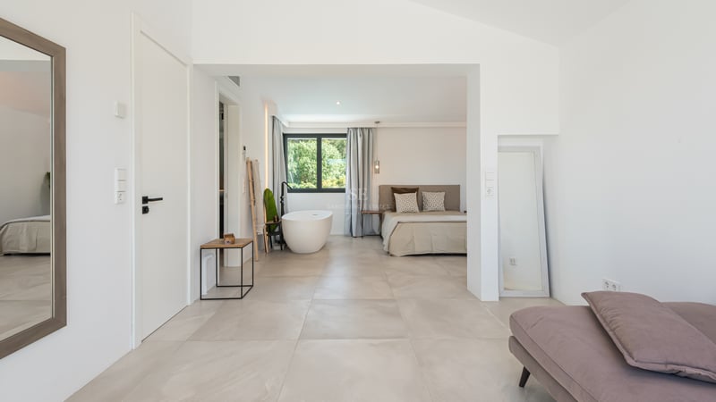 Chambre principale lumineuse avec carrelage grand format, lit minimaliste et baignoire îlot blanche près d'une fenêtre.