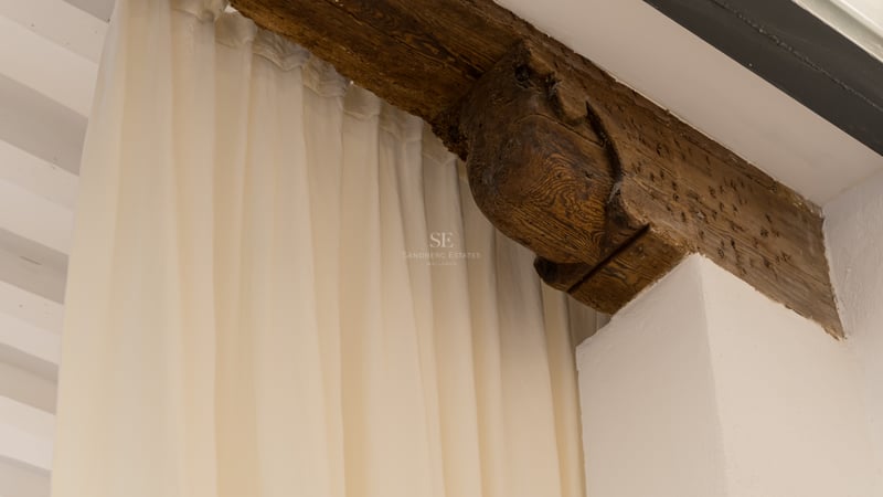 Poutre en bois sombre sculptée à la main à côté de rideaux crème et mur blanc.