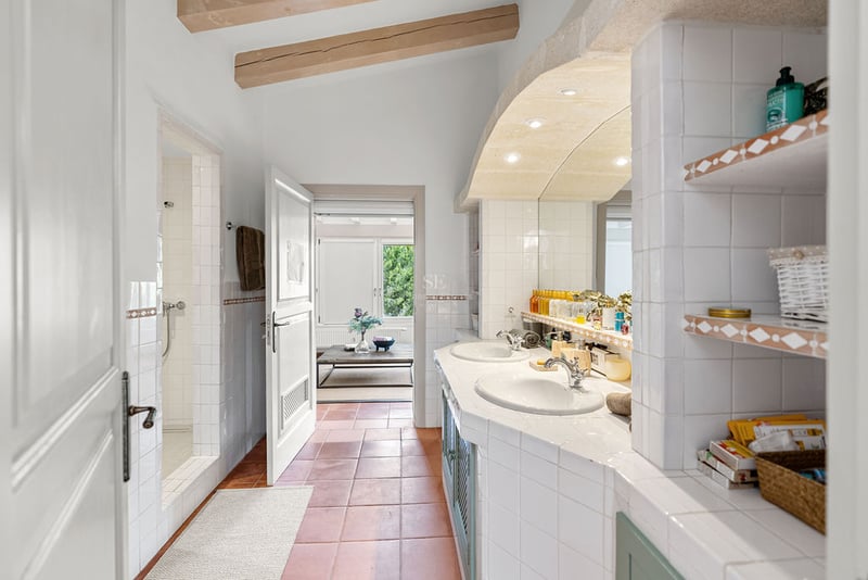 Salle de bain spacieuse avec sol en terre cuite, double vasque blanche, poutres en bois et douche à l'italienne.