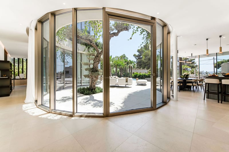 Intérieur d'une villa de luxe avec mur de verre incurvé donnant sur un patio privé avec olivier.