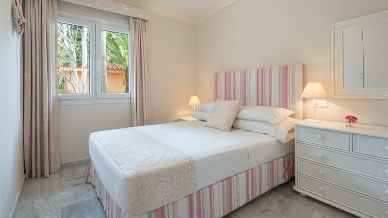 Chambre avec lit double, textiles roses, sol en marbre blanc et fenêtre donnant sur des palmiers.