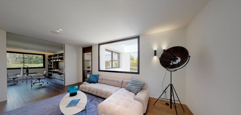 Salon moderne avec canapé capitonné crème, tapis bleu et grande fenêtre donnant sur un patio ensoleillé.