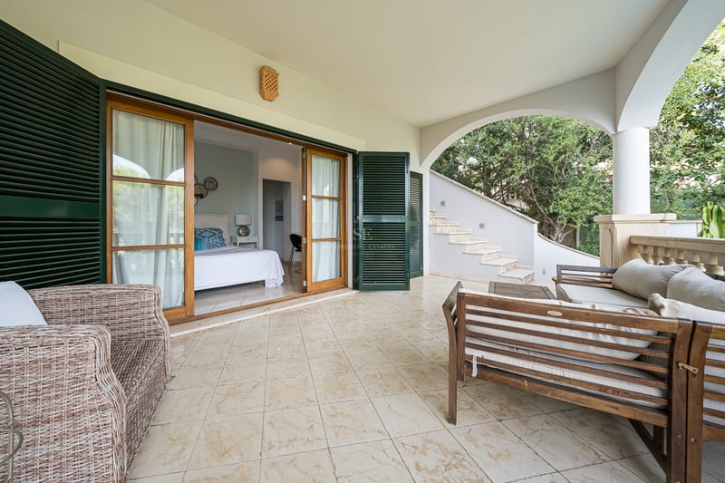 Terrasse méditerranéenne couverte avec carrelage, meubles en bois et grandes baies vitrées menant à une chambre.