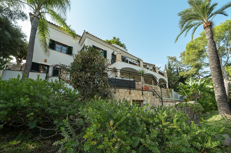Villa blanche de trois étages avec terrasses voûtées, murs en pierre et palmiers sous un ciel bleu.