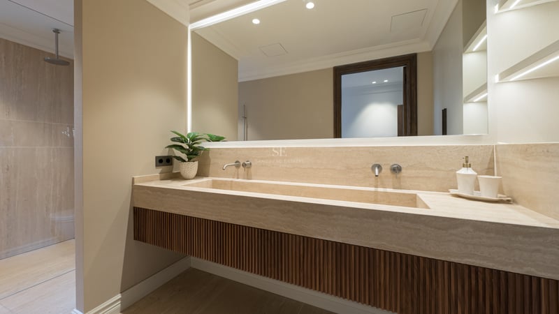 Luxury bathroom featuring a custom travertine integrated sink, fluted wood cabinetry, and a large backlit mirror.