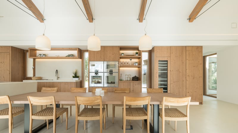 Light wood dining table with woven chairs in front of a modern oak kitchen featuring integrated appliances and wine cooler.