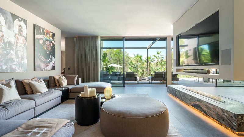 Salon moderne avec canapé d'angle gris, cheminée linéaire et baies vitrées ouvrant sur un jardin tropical.
