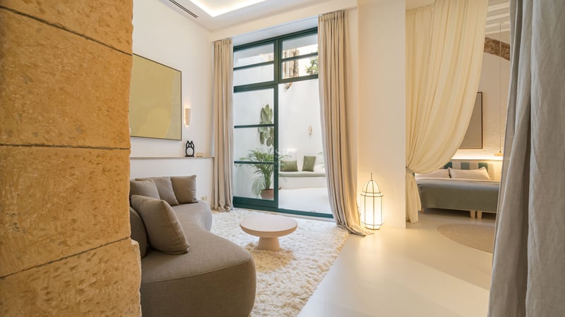 Salon lumineux avec canapé beige incurvé, tapis blanc moelleux et grandes portes vitrées ouvrant sur un patio intérieur.