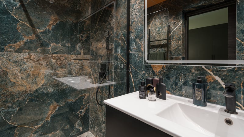 Salle de bains haut de gamme avec murs en marbre vert foncé, veines dorées, vasque blanche et robinetterie noire mate.