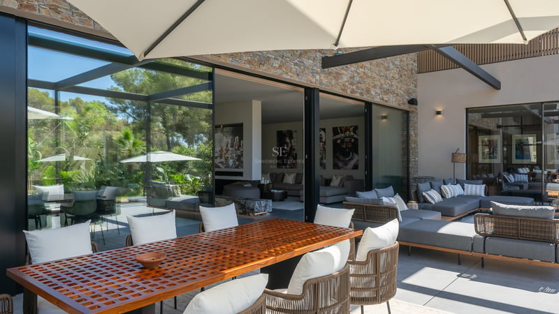 Terrasse moderne avec une grande table en bois, des canapés gris et des baies vitrées ouvrant sur le salon.